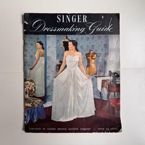 Op de afbeelding: Een vintage Singer Dressmaking Guide boekje met een omslagillustratie van een persoon in een witte strapless japon in een kamer met een paspop, een naaimachine op een tafel en een bloemrijke stoel. De tekst op de omslag is Singer Dressmaking Guide.