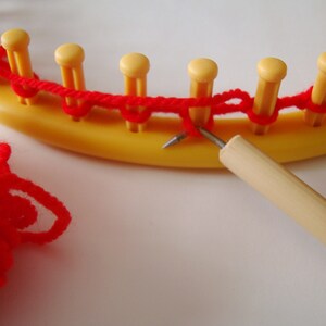 May include: A close-up of a red yarn being used to knit on a yellow circular loom. A wooden knitting needle is being used to pull the yarn through the loops.