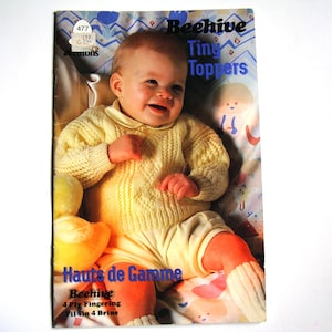 May include: A knitting pattern booklet for a baby sweater and booties. The cover features a photo of a baby wearing a yellow knitted sweater and booties. The title of the pattern is "Beehive Tiny Toppers" and the subtitle is "Hauts de Gamme".