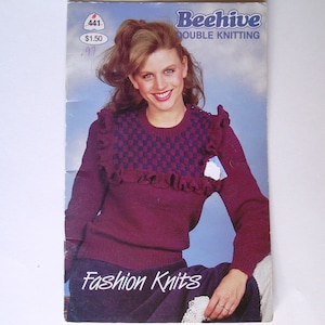 Könnte beinhalten: Ein Vintage-Strickmusterheft mit einer Frau, die einen burgunderfarbenen Pullover mit einem blauen und burgunderfarbenen gemusterten Joch und einem Rüschenkragen trägt. Das Cover lautet "Beehive Double Knitting Fashion Knits" und hat ein Preisschild von 1,50 $.