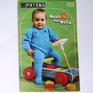 Könnte beinhalten: Ein Vintage-Strickmusterbuchcover mit einem Baby in einem blauen Strickoverall, das auf einem Spielzeugwagen mit bunten Blöcken sitzt. Der Buchtitel lautet "Beehive for Baby" und der Verlag ist Patons.