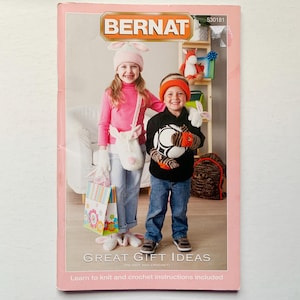Op de afbeelding: Een roze en wit boek met de titel "Bernat Great Gift Ideas" en de tekst "Learn to knit and crochet instructions included". De omslag toont twee kinderen die gebreide mutsen dragen en gebreide speelgoed vasthouden.