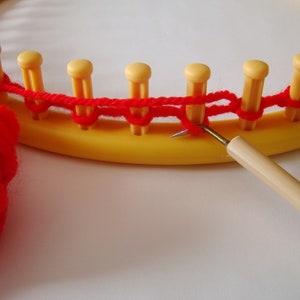 Peut inclure: Gros plan sur un fil rouge utilisé pour tricoter sur un métier à tisser circulaire jaune. Une aiguille à tricoter en bois est utilisée pour tirer le fil à travers les boucles.