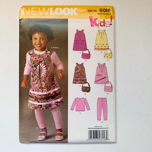 Op de afbeelding: Een naaipatroon voor kinderkleding, met een roze en bruine jurk met bloemenprint, een roze top en een roze broek. Het patroon is van New Look, bin 70, 6016, maat 1/2-4.