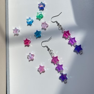 Op de afbeelding: Oorbellen met stervormige kralen in roze, paarse en blauwe tinten. De oorbellen hangen aan zilverkleurige haken. Verspreid rond de oorbellen liggen extra stervormige kralen in verschillende kleuren. De achtergrond is wit.