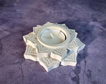 Puede incluir: Un portavelas de hormigón gris con forma de flor de loto. Tiene una hendidura circular para sostener una pequeña vela blanca. El diseño de la flor de loto es visible en los bordes.