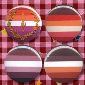 May include: Four circular pin-back buttons featuring various horizontal striped patterns. One button displays a central gold sword emblem surrounded by a gold laurel wreath over a multi-colored striped background of orange, white, and purple.