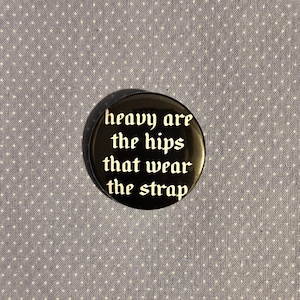 Puede incluir: Un botón negro con la frase "heavy are the hips that wear the strap" en blanco, con letras góticas. El botón está sobre un fondo de tela azul claro con un patrón de lunares blancos.