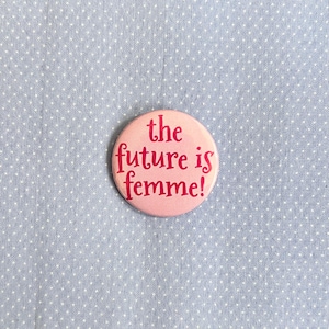 Może przedstawiać: Okrągła, różowa przypinka z napisem "the future is femme!" w czerwonym kolorze. Przypinka umieszczona na jasnoniebieskiej tkaninie w białe grochy.
