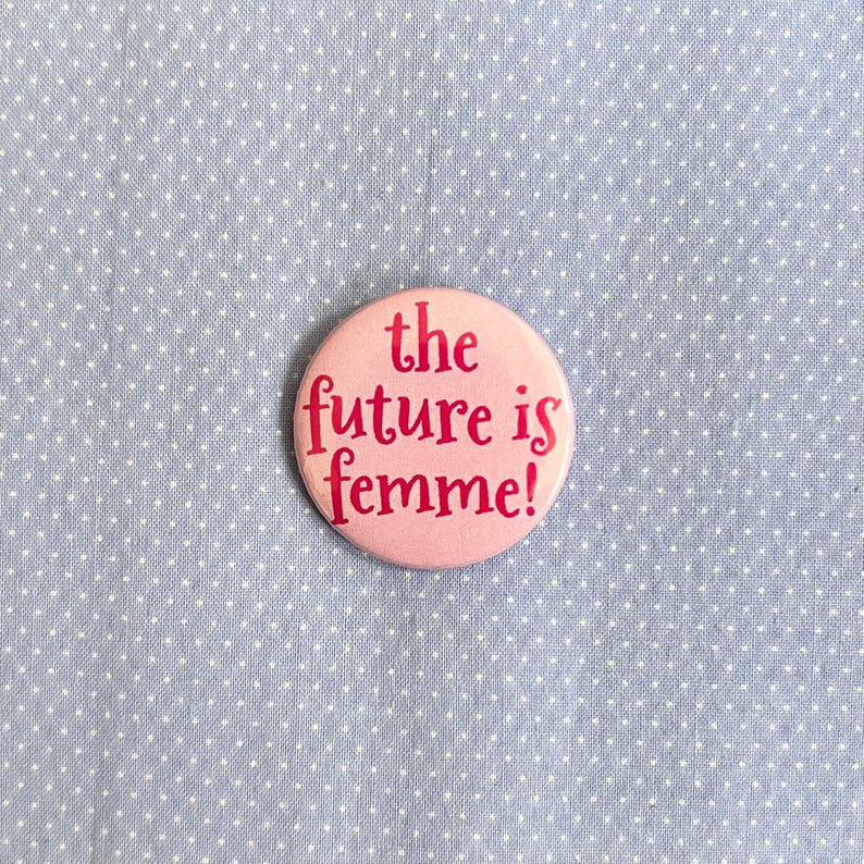 以下が含まれることがあります： 「the future is femme!」の赤い文字が書かれた丸いピンク色のバッジ。白い水玉模様の入った水色の生地の上に配置されています。
