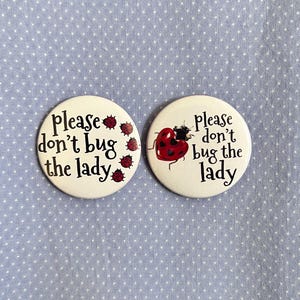 Op de afbeelding: Twee witte ronde knopen met de tekst "Please don't bug the lady." Eén knoop heeft een lieveheersbeestje-illustratie, de andere heeft lieveheersbeestje-illustraties. De knopen liggen op een lichtblauwe gestippelde achtergrond.