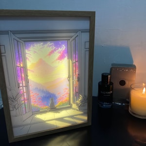 Puede incluir: Una ilustración enmarcada de un gato mirando por una ventana a un paisaje de puesta de sol colorido. La ventana está abierta y el gato está sentado en el alféizar de la ventana. El marco es de madera y tiene un acabado marrón claro.