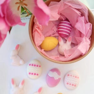 May include: A collection of pastel pink and yellow Easter eggs and white bunny figurines. The eggs are decorated with white and pink stripes, white dots, and a wavy pattern. The bunnies are in various poses, including sitting and standing.
