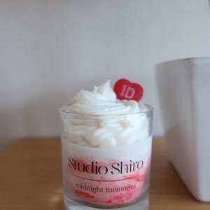 May include: A clear glass jar candle with a white wax top and a red heart-shaped topper with the number "1D" on it. The candle has a pink and red ombre base with the text "Studio Shiro" and "midnight memories" on the label.