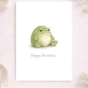 May include: A greeting card with a watercolor illustration of a green frog with a grumpy expression. The frog is sitting down with its arms and legs at its sides. The text "Hoppy Birthday" is printed below the frog.