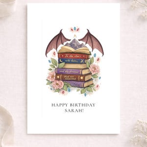 May include: A greeting card featuring a stack of books with bat wings, a mountain peak, and pink flowers. The books are inscribed with the text: To the stars who listen and the dreams that are answered. Below the illustration is the text: HAPPY BIRTHDAY SARAH!