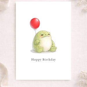 May include: A white greeting card featuring a cartoon frog holding a red balloon. The frog is light green with a slightly grumpy expression. The text "Hoppy Birthday" is printed below the frog.