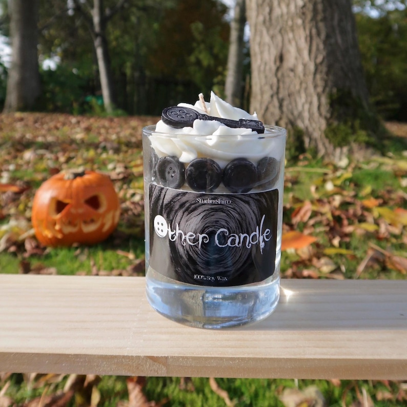May include: A white candle with black buttons and a key on top. The candle is in a clear glass jar with a black label that says "Other Candle" and "100% Soy Wax".