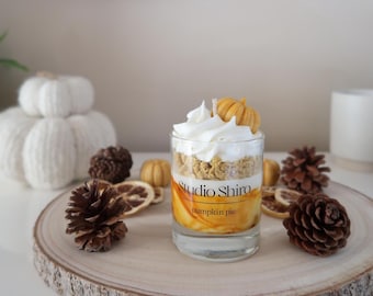 Op de afbeelding: Een glazen kaars pot met een pumpkin pie geur kaars. De kaars is bedekt met een witte slagroom frosting en een kleine oranje pompoen. De pot heeft een zwart label met de tekst "Studio Shiro pumpkin pie".