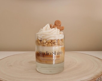 May include: A clear glass jar candle with a white frosting-like top, brown sprinkles, and a brown candy on top. The candle is labeled "Studio Shiro" and "freshly baked gingerbread". The candle is sitting on a wooden slice.