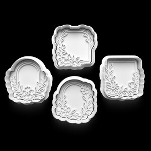 Op de afbeelding: Vier witte plastic koekjessnijders in verschillende vormen: twee boogvormige, een vierkante en een rechthoekige. Elke snijder heeft een bloemen- en bladerontwerp langs de randen, waardoor een decoratieve rand ontstaat. Op een zwarte achtergrond.