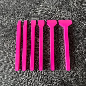 Op de afbeelding: Zes felroze plastic gereedschappen van verschillende vormen en maten zijn gerangschikt op een getextureerd donkergrijs oppervlak. De gereedschappen lijken te zijn voor knutselen of modelleren, met platte, spatelachtige uiteinden.