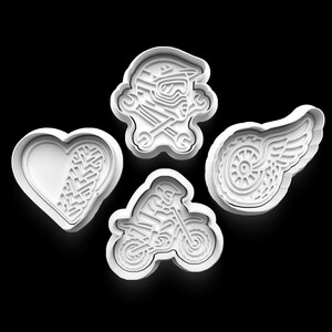 Op de afbeelding: Vier witte koekjessnijders in verschillende vormen. Eén is een hart met bandensporen, een andere is een motorhelm met gekruiste sleutels, een derde is een motorfiets met een rijder en de laatste is een wiel met vleugels.
