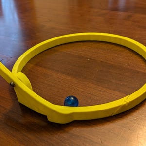 Pode incluir: Um aro de plástico amarelo com uma alavanca lateral. Uma bola de gude azul está dentro do aro.