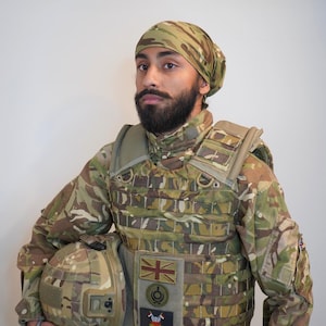 Op de afbeelding: Een man in een camouflage uniform met een zwarte bandana, een vest met een Britse vlag patch en een helm.