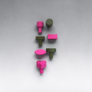 May include: An assortment of colorful, textured objects in shades of pink and green. The objects have various shapes, including cylindrical, rectangular, and oval, with a T-shaped base. The objects are arranged on a light gray background.