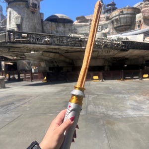 May include: A long, golden brown churro is held up like a lightsaber in front of a Star Wars themed background. The churro is decorated with gold and silver accents, resembling a lightsaber.