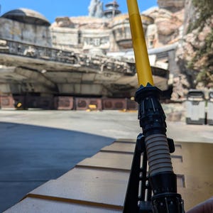 May include: A yellow lightsaber with a black handle and a black stand. The lightsaber is on a yellow surface.