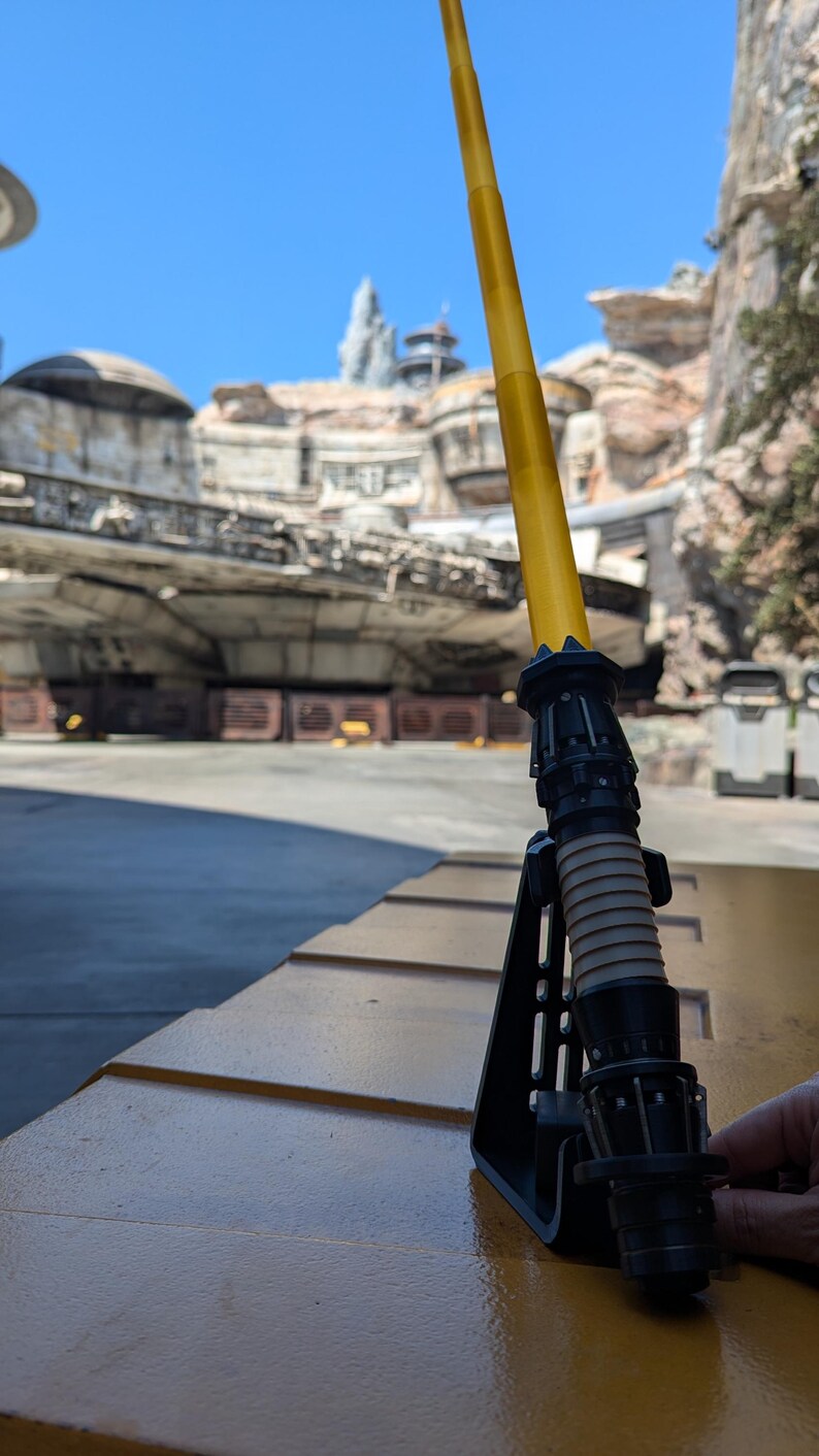 May include: A yellow lightsaber with a black handle and a black stand. The lightsaber is on a yellow surface.