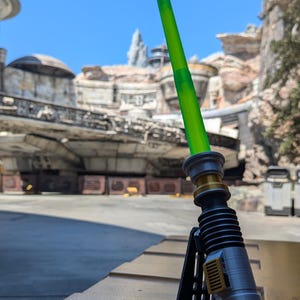 May include: A green lightsaber with a silver handle and a black stand. The lightsaber is on a yellow surface in front of a building.