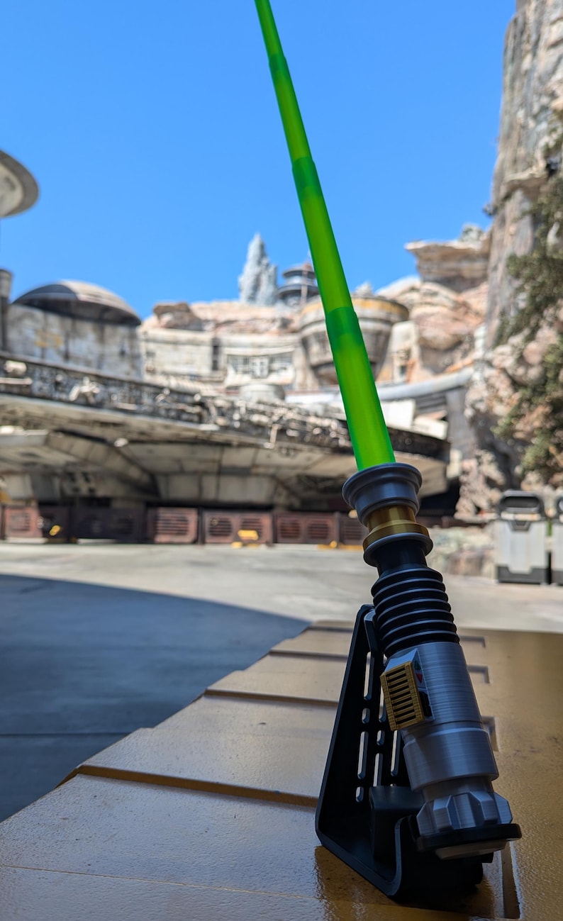May include: A green lightsaber with a silver handle and a black stand. The lightsaber is on a yellow surface in front of a building.