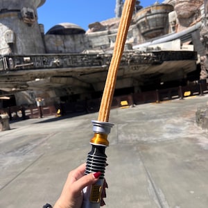 May include: A long, golden brown churro is held in a hand, resembling a lightsaber. The churro is decorated with a silver and gold lightsaber handle. The background features a large, gray spaceship-like structure.