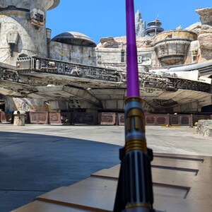 May include: A purple lightsaber with a gold hilt is standing upright on a wooden surface. The lightsaber is in front of a Star Wars themed building and a spaceship.