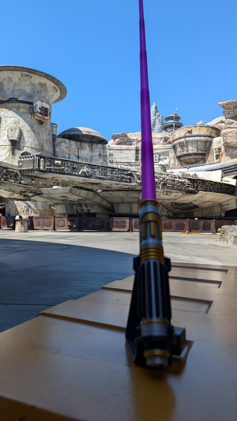 May include: A purple lightsaber with a gold hilt is standing upright on a wooden surface. The lightsaber is in front of a Star Wars themed building and a spaceship.