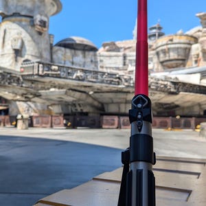 May include: A red lightsaber with a black handle and a silver blade is standing on a yellow surface. The lightsaber is in a stand. The background is a blurry image of a Star Wars themed park.