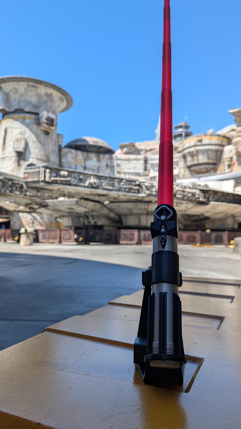 May include: A red lightsaber with a black handle and a silver blade is standing on a yellow surface. The lightsaber is in a stand. The background is a blurry image of a Star Wars themed park.
