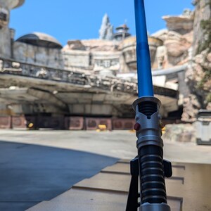 May include: A blue lightsaber with a silver handle and black accents is displayed on a black stand. The lightsaber is in a retracted position. The background is a blurry image of a Star Wars themed amusement park.