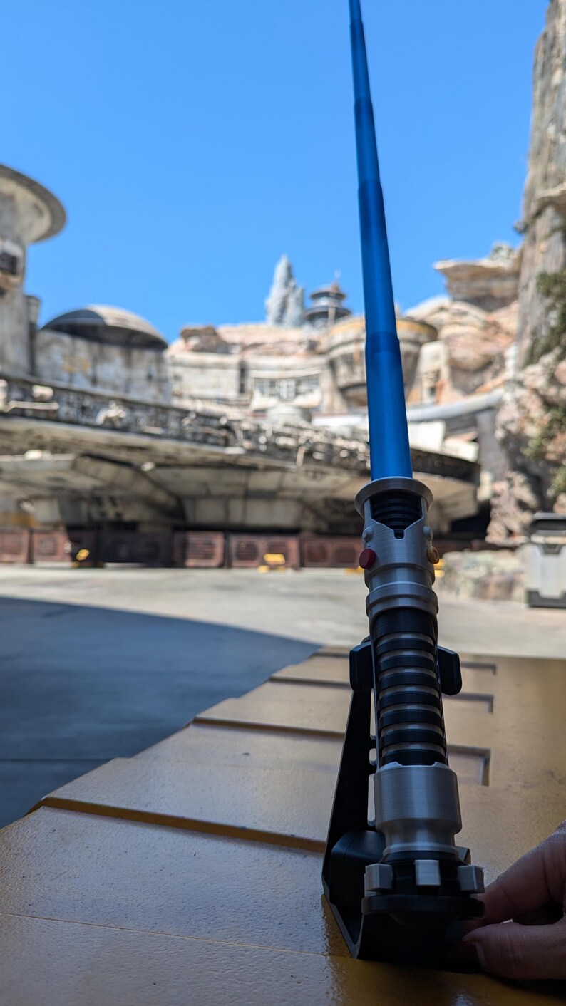 May include: A blue lightsaber with a silver handle and black accents is displayed on a black stand. The lightsaber is in a retracted position. The background is a blurry image of a Star Wars themed amusement park.