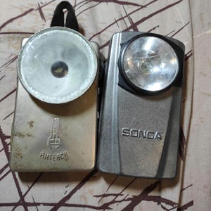 May include: Two vintage flashlights. One is silver with a large circular light and the word "Витебск" printed on it. The other is grey with the word "SONGA" printed on it and a smaller circular light.