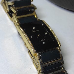 May include: A Rado Jubilé wristwatch featuring a rectangular black dial with four small diamond hour markers, gold-tone hands, and a bezel encrusted with small clear stones. The bracelet consists of alternating black ceramic and gold-tone metal links.