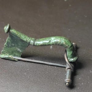 May include: An antique bronze brooch with a green patina. The brooch features a curved bar, a pin, and a decorative element. The metal has a weathered appearance, suggesting age. The brooch is likely a vintage or antique jewellery piece.