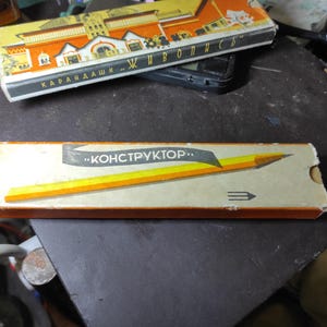 May include: A vintage pencil box with the word "КОНСТРУКТОР" in a banner, featuring a yellow and orange pencil illustration. The box is rectangular with a light beige background and a brown border. Another box with Russian text is visible.