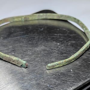Könnte beinhalten: Ein antikes Bronze-Armband mit verwitterter, grünspanfarbener Oberfläche. Das Armband ist offen, mit einem sichtbaren Bruch an einem Ende. Das Metall hat eine strukturierte Oberfläche, die auf Alter und Gebrauch hindeutet. Das Armband wird auf einer metallischen Oberfläche präsentiert.