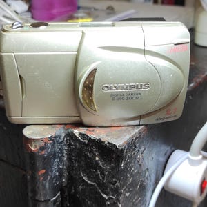 May include: A silver Olympus C-990 Zoom digital camera with a 2.1 megapixel resolution. The camera has a built-in flash and a zoom lens. The word "CAMEDIA" is written in pink on the top right corner.