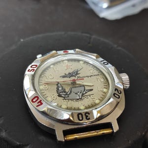 May include: A silver-toned wristwatch with a beige dial featuring a graphic of a jet and a ship. The dial includes a red star, Cyrillic text, and luminous hour markers. The rotating bezel has black numerals 10, 20, 30, 40, and 50.