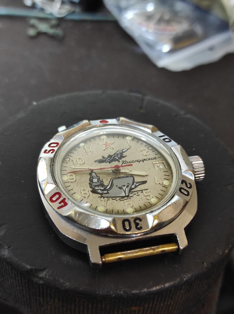 May include: A silver-toned wristwatch with a beige dial featuring a graphic of a jet and a ship. The dial includes a red star, Cyrillic text, and luminous hour markers. The rotating bezel has black numerals 10, 20, 30, 40, and 50.
